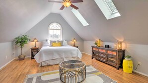 A bedroom with a sloped ceiling, a bed with white bedding, two bedside lamps, a wooden dresser, a round wicker ottoman, and a skylight.