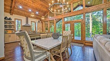 Interior de madera con una mesa y sillas de comedor, un candelabro y grandes ventanales con vistas al bosque.