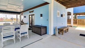 Un espace extérieur abrité avec des chaises blanches et un banc en bois, un meuble de rangement sombre et un bâtiment bleu en arrière-plan.