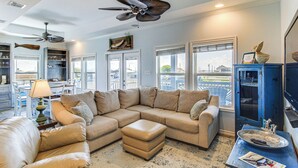 Un salon avec un canapé d’angle, un pouf, un ventilateur de plafond et une vue dégagée grâce à de grandes fenêtres.