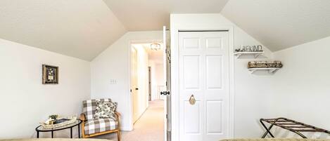 A cozy bedroom with a checkered armchair, a small table with a basket, a white door, and a shelf with decorative items.