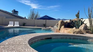 A swimming pool with a curved edge, surrounded by a stone deck. There are two lounge chairs and a blue umbrella. - Lake Havasu City House w/ Fire Pit, Pool & Spa! (Lake Havasu City)