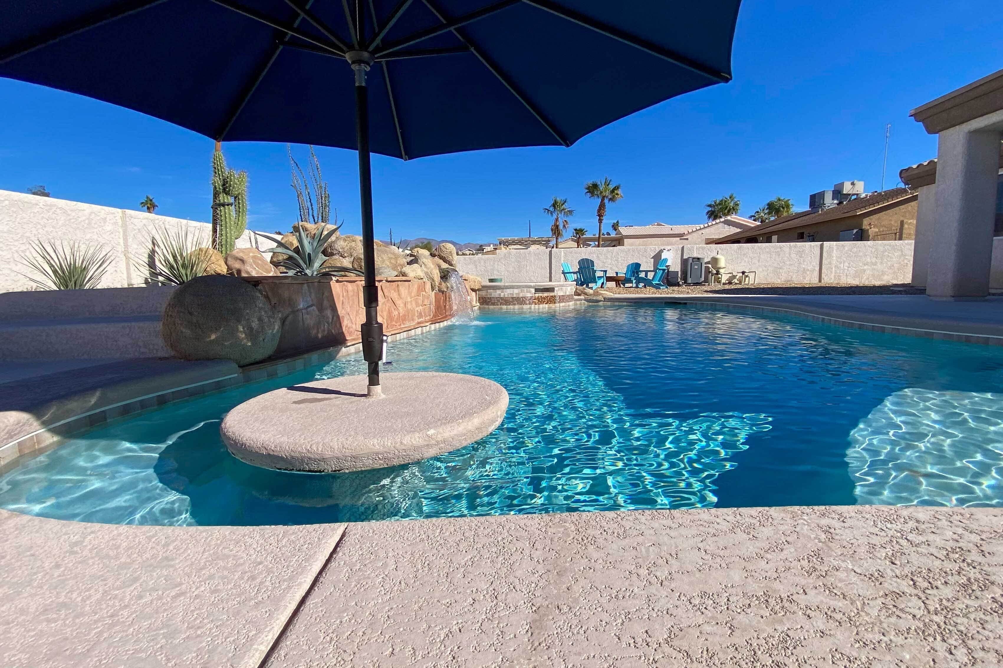 A swimming pool with clear blue water, surrounded by a stone deck, and a large umbrella providing shade.