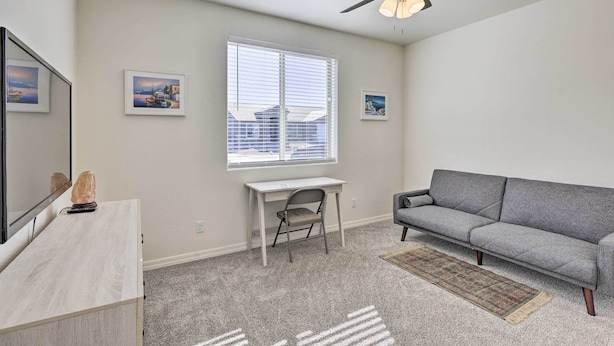 A living room with a grey sofa, a white desk, a chair, a ceiling fan, and a window with blinds. - Modern Kingman Vacation Rental, 5 Mi to Dtwn! (Kingman)