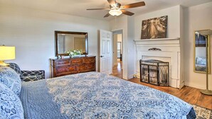 A bedroom with a large bed, a ceiling fan, a dresser, a mirror, a fireplace, and a door leading to another room. - Deck, Grill & Yard: Pet-friendly Van Alstyne Gem! (Van Alstyne)