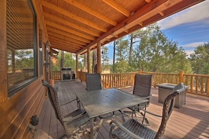 A covered outdoor deck with a table and chairs, a grill, and a view of trees.