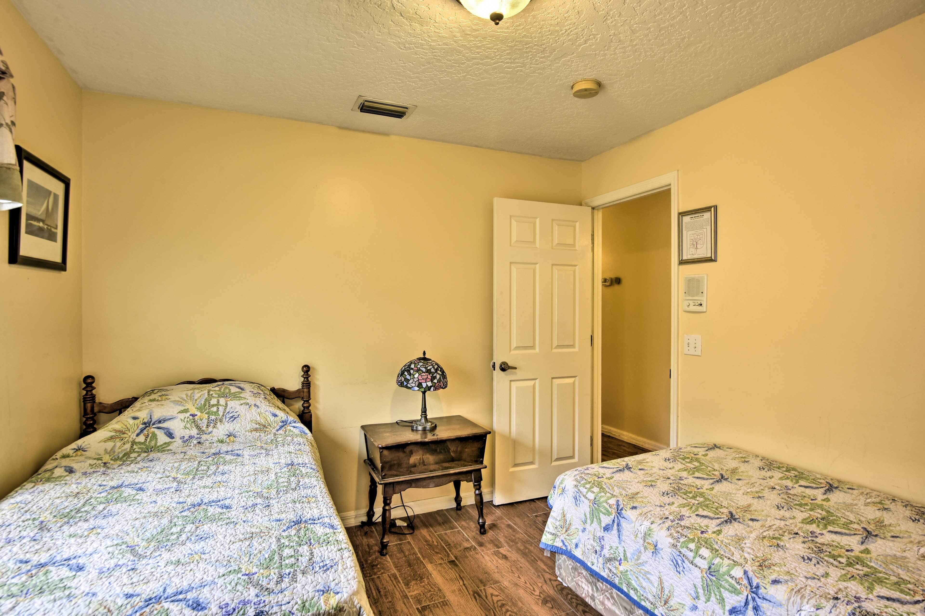 A bedroom with two beds, a nightstand with a lamp, a door, and a wall-mounted light fixture.