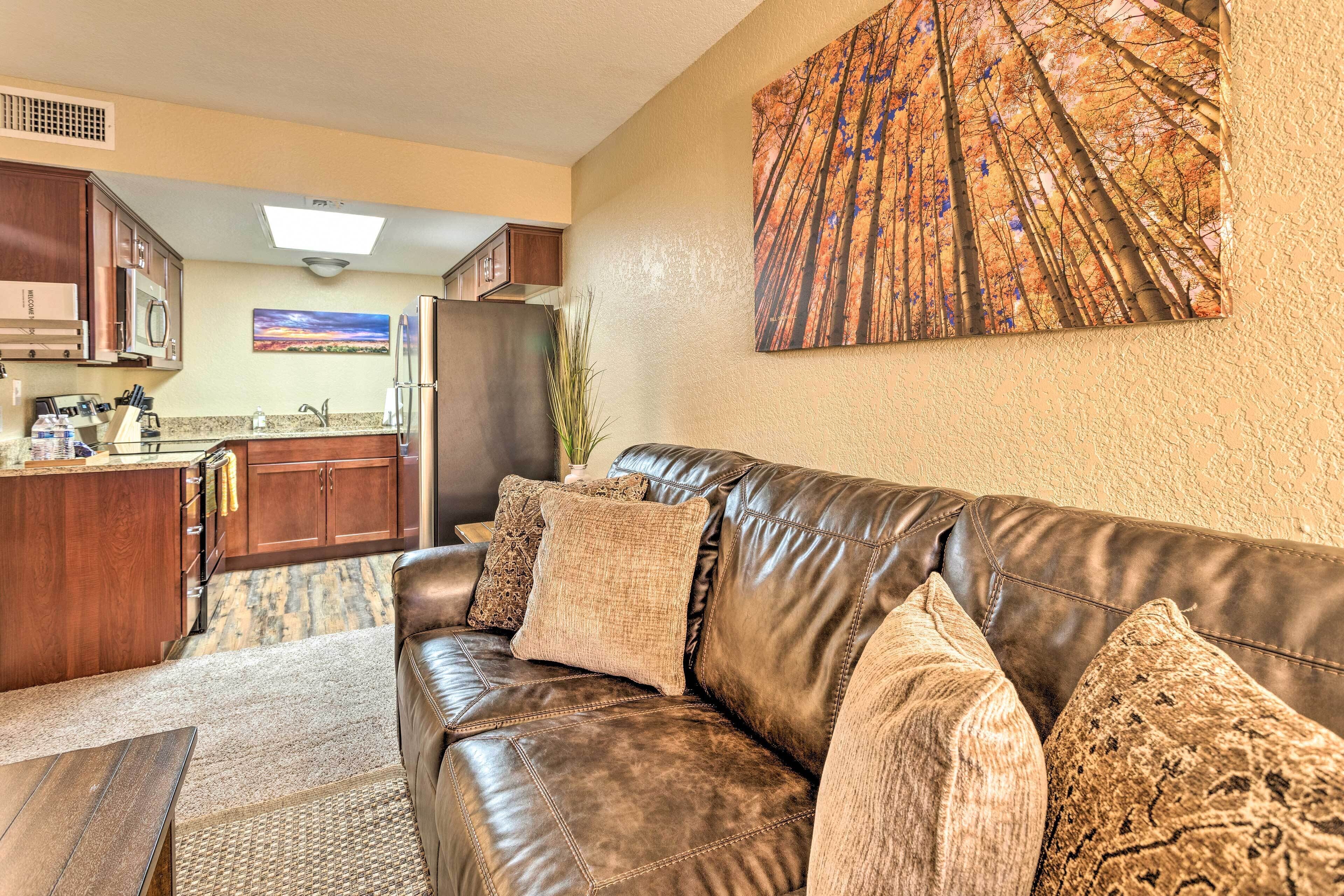 A living room with a brown leather couch, a kitchen in the background, and a large painting on the wall.