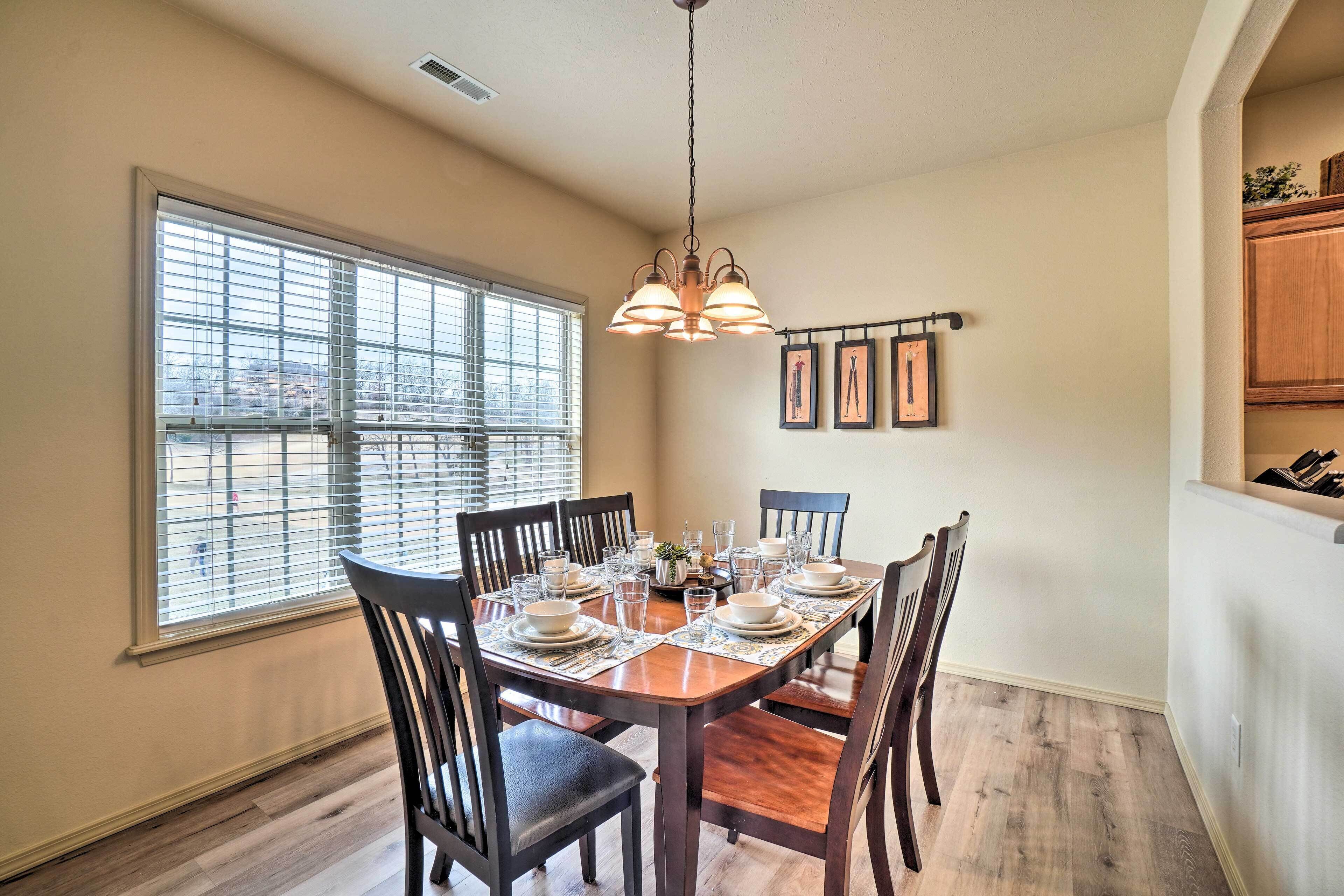 A dining area with a wooden table set for four, surrounded by chairs, with a large window and a pendant light above.