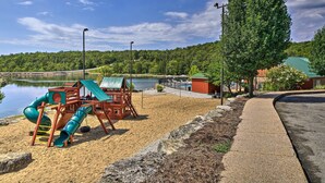 A playground with a slide and climbing structure near a lake, surrounded by trees and a paved path. - Branson West Villa w/ Golf Course View & Pool! (Branson West)