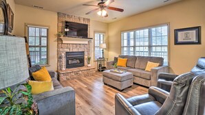 A living room with a stone fireplace, a ceiling fan, and large windows. - Branson West Villa w/ Golf Course View & Pool! (Branson West)