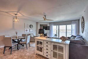 A modern living room with a dining area, a ceiling fan, a large window, and a view of the outdoors.