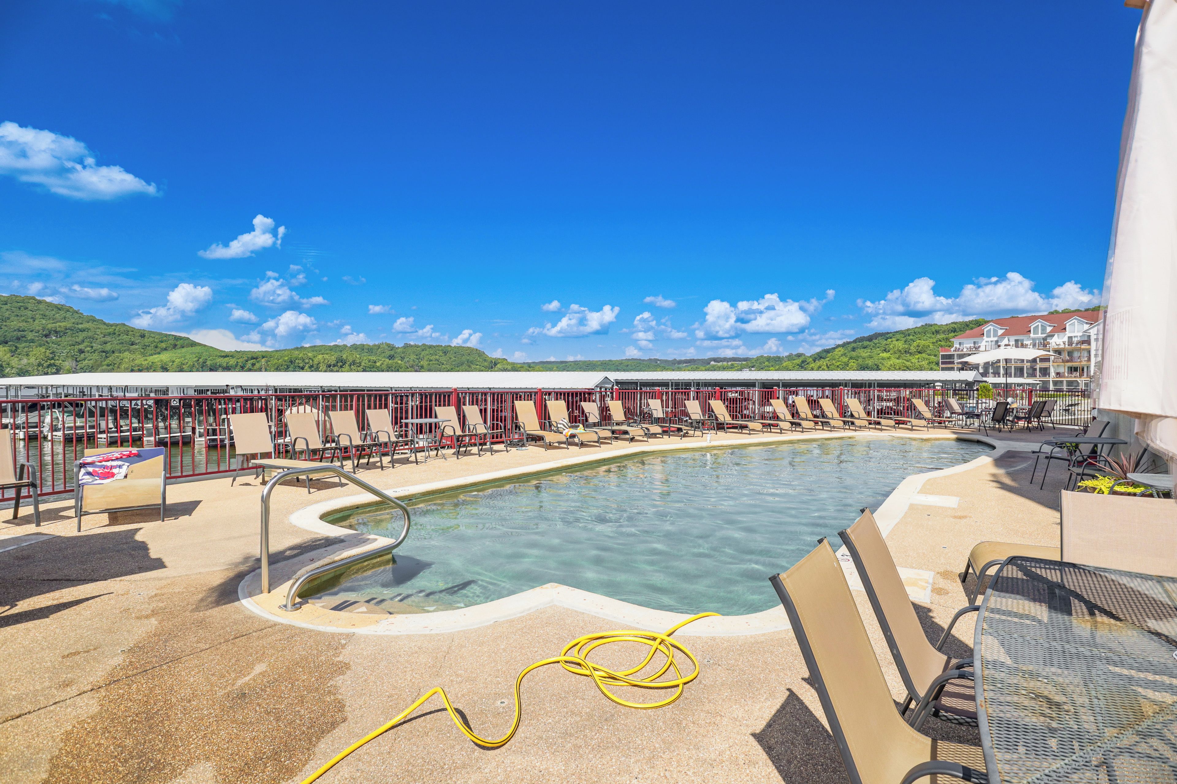 Un espace piscine avec des chaises longues et une table placée au bord de la piscine, le tout entouré d’une clôture rouge et, en arrière-plan, d’un immeuble doté de nombreux balcons.