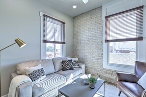 A modern living room with a grey sofa, a coffee table, and a brown leather armchair. There are two windows with blinds, a brick wall, and a floor lamp. - Bright San Marcos Apartment w/ Ideal Location (San Marcos)