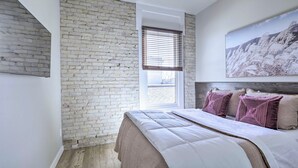 A bedroom with a brick wall, a window with blinds, a bed with white bedding, and a painting on the wall. - Bright San Marcos Apartment w/ Ideal Location (San Marcos)