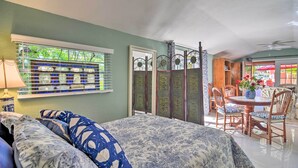 A bedroom with a bed, a window with blinds, a bedside table with a lamp, a dining area with chairs and a table, and a view of the outside.