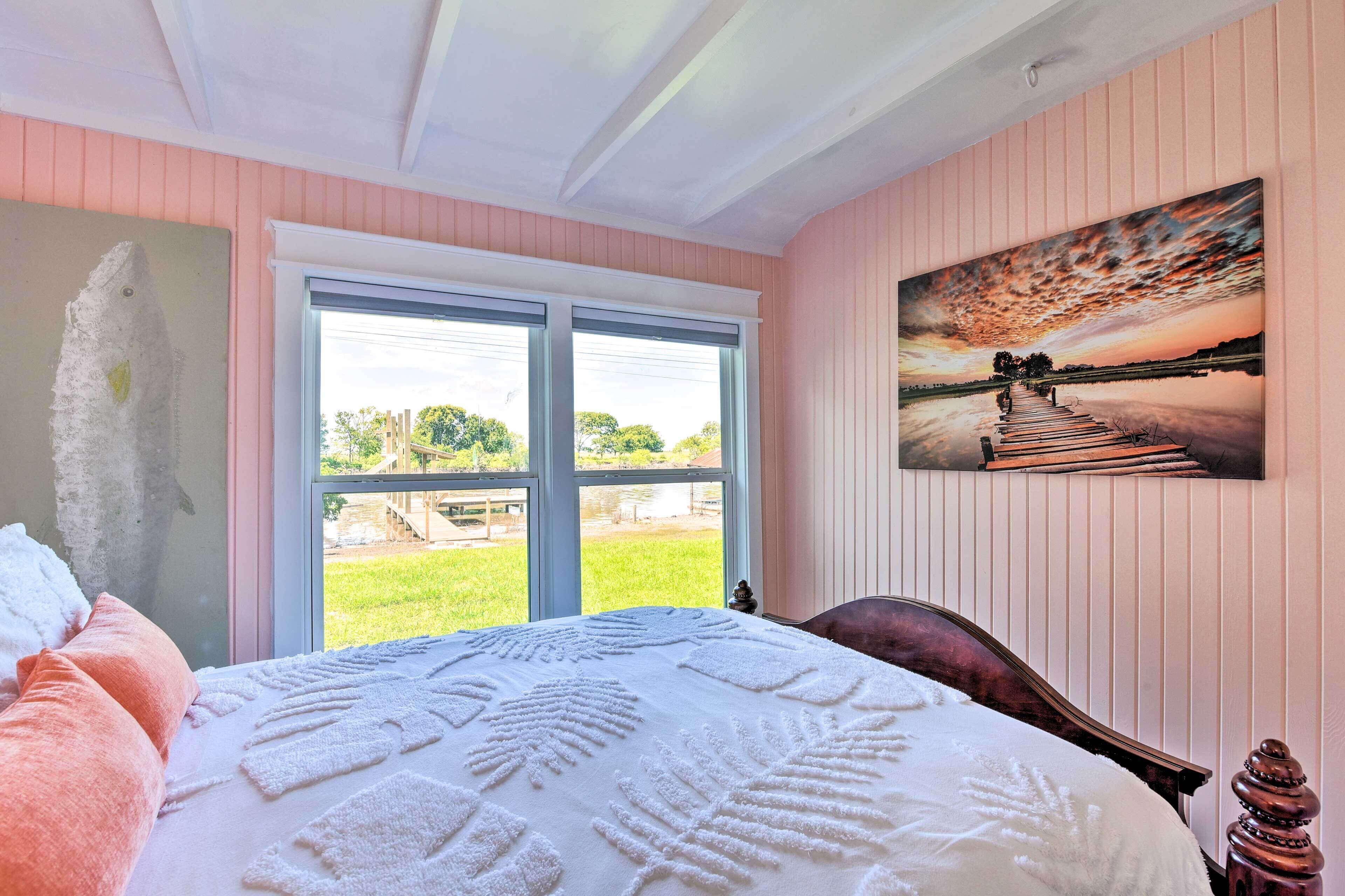 A bedroom with a large bed, a window with a view of a grassy area, and a painting of a sunset on the wall.