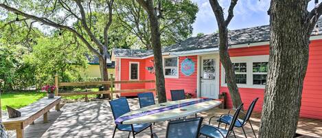 A wooden deck with a table and chairs, surrounded by trees and a red building with a white door.