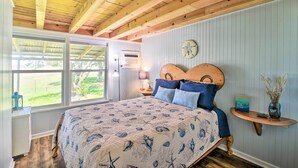 A bedroom with a wooden ceiling, a bed with a patterned bedspread, a nightstand with a lamp, and a window with a view of greenery. - Furnished Deck & Dock: Renovated Sargent Cabin! (Bay City)