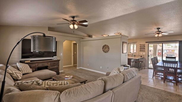 A spacious living room with a large sectional sofa, a flat-screen TV, and ceiling fans. There is a dining area with a table and chairs visible in the background.