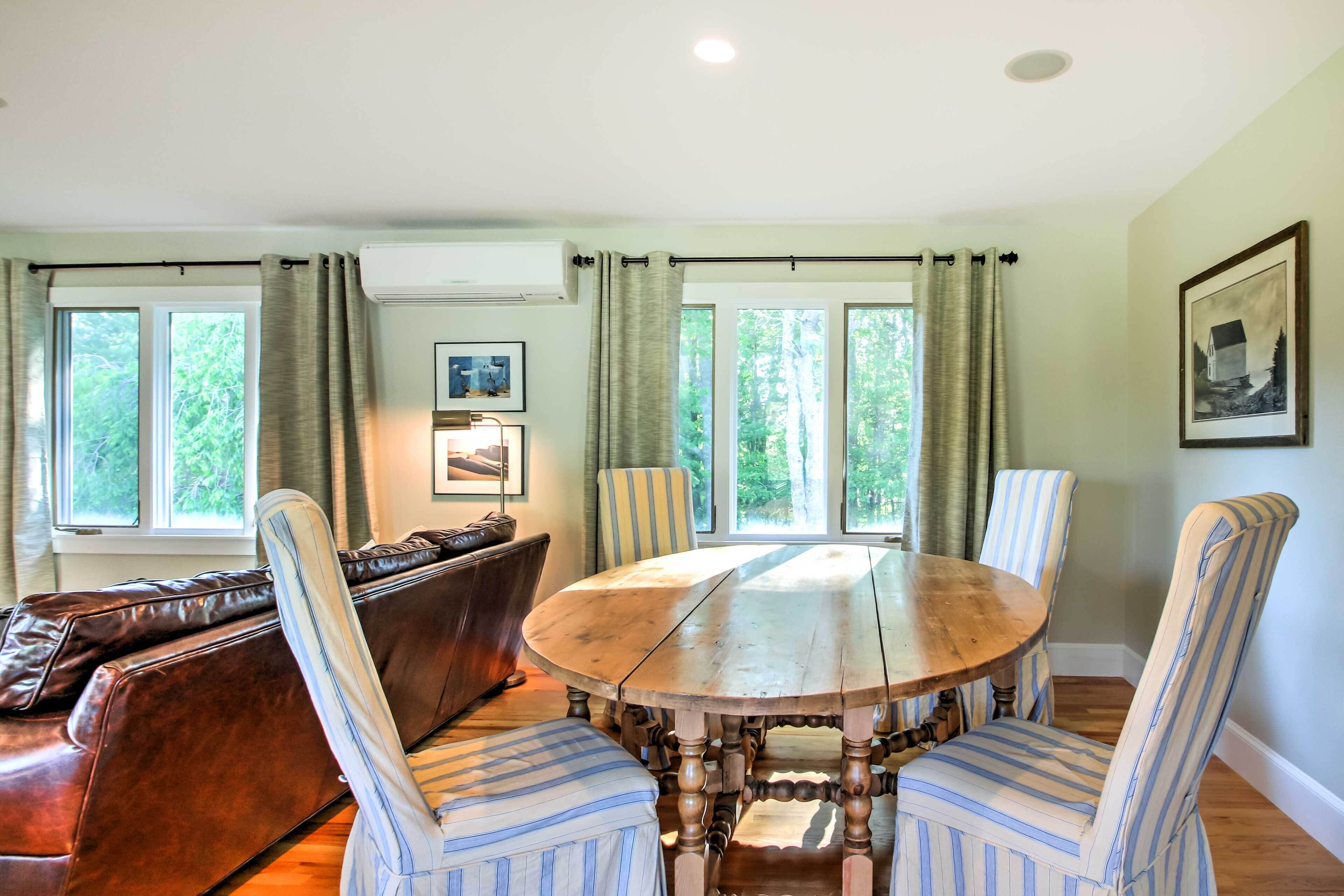 Uma sala de jantar com mesa de madeira e cadeiras listradas, um sofá de couro marrom e uma janela com cortinas.