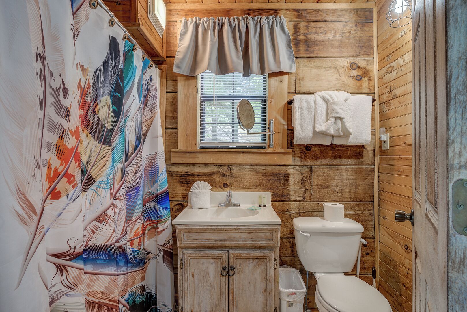 Un baño rústico con paredes de madera, cortina de ducha con estampado de pájaros, una ventana con persianas, un lavabo con artículos de aseo y un inodoro.
