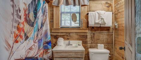 Un baño rústico con paredes de madera, cortina de ducha con estampado de pájaros, una ventana con persianas, un lavabo con artículos de aseo y un inodoro.