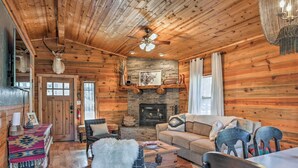 A log cabin interior with a fireplace, wooden walls, a ceiling fan, and rustic furnishings. - Ruidoso Log Cabin Retreat - Peaceful & Convenient (Ruidoso)