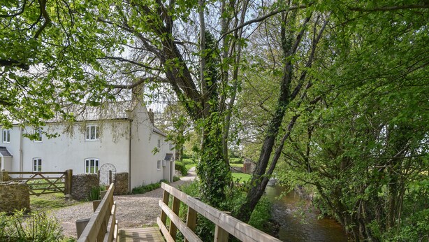 A wooden bridge leading to a white building with a stone wall and a metal gate. - Millwater Cottage (Axminster)