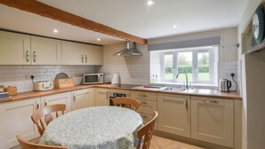 A kitchen with wooden cabinets, a round table with chairs, a window with a view of greenery, and a kettle on the counter. - Millwater Cottage (Axminster)