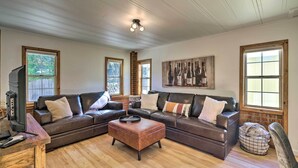 A living room with a brown leather sectional sofa, a wooden coffee table, and a television on a wooden table. There are windows with wooden frames and a brick wall. - Wine Country Escape w/ Hot Tub in River Pines! (River Pines)