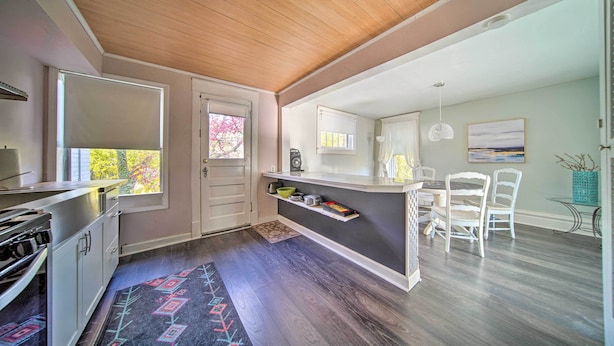 Een moderne keuken met een houten plafond, een eethoek met witte stoelen en uitzicht op bomen door een raam.