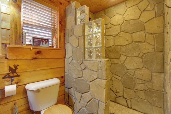 Une salle de bain rustique, située dans une cabane en bois, avec un mur en pierre, des toilettes et une fenêtre équipée de stores.