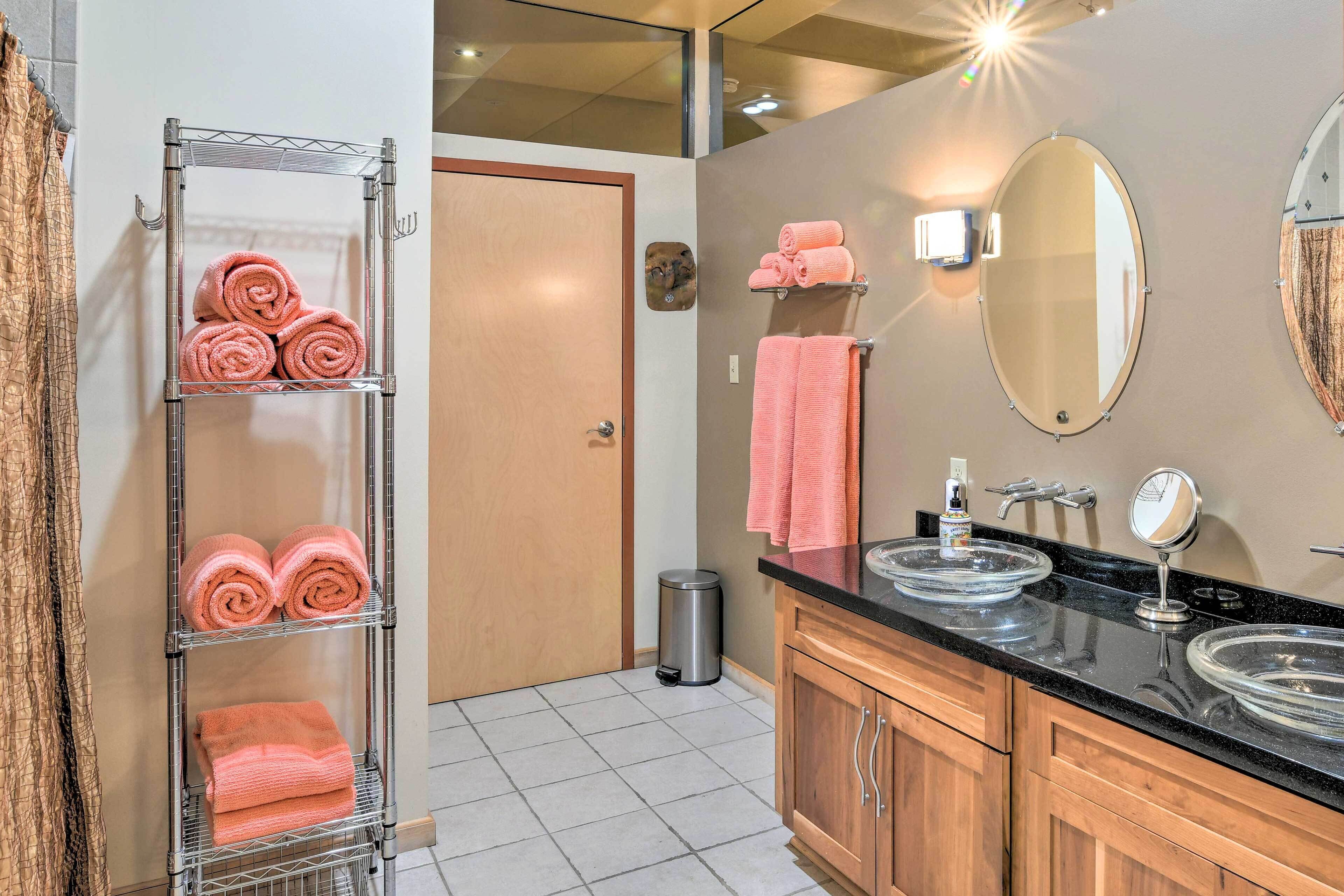 A bathroom with a metal towel rack holding pink towels, a wooden vanity with two sinks, a mirror, and a trash can.