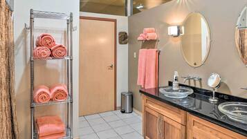A bathroom with a metal towel rack holding pink towels, a wooden vanity with two sinks, a mirror, and a trash can.