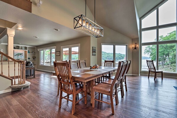 A spacious dining area with a large wooden table and chairs, a modern chandelier, and large windows offering a view of the outdoors. - Lakefront Haven: Family-friendly Sunrise Beach Gem (Sunrise Beach)