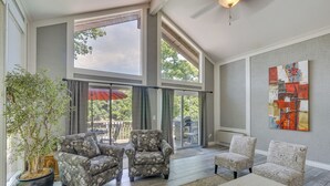 A spacious living room with large windows, a potted plant, and a coffee table with remotes.