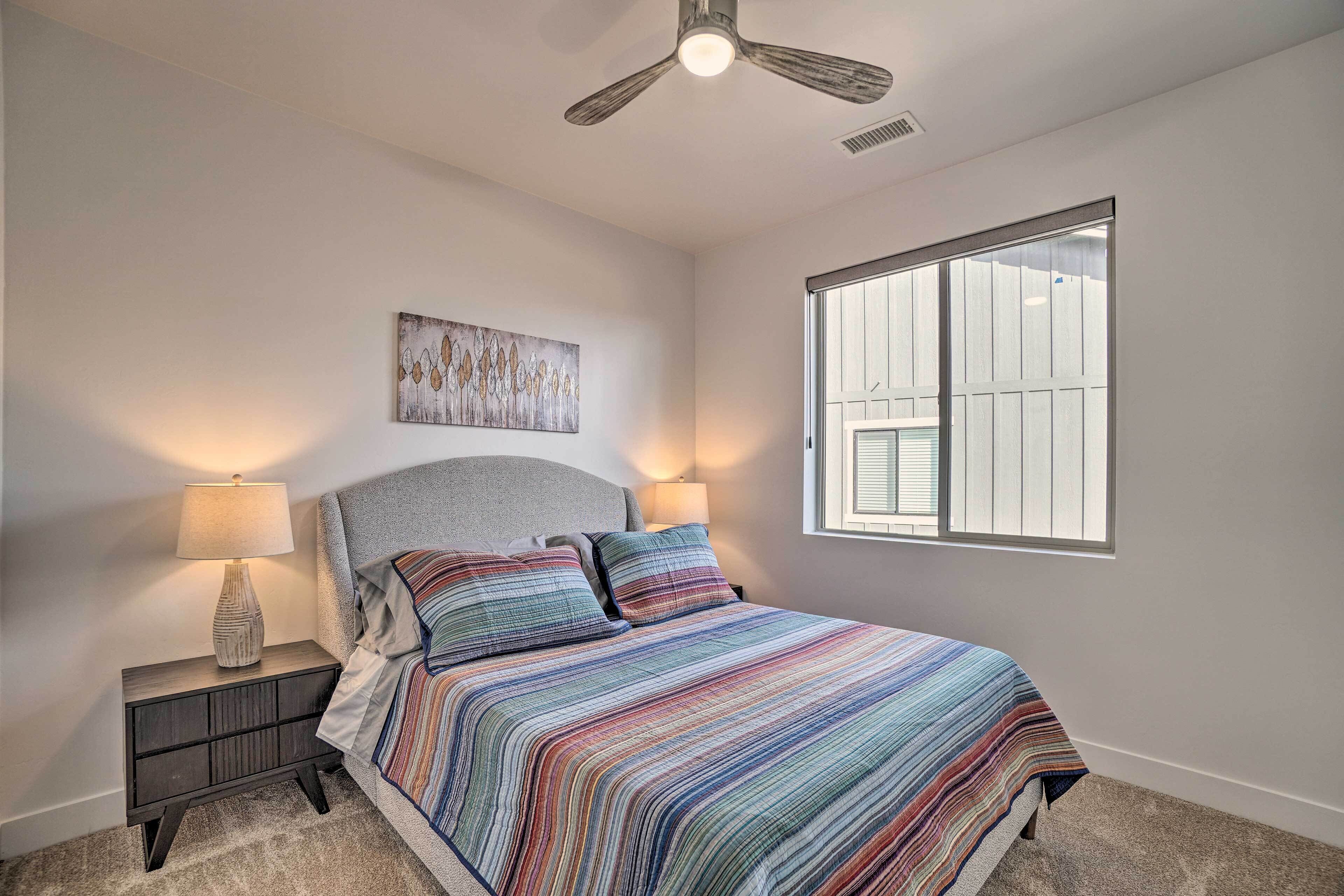 A bedroom with a bed, two bedside lamps, a ceiling fan, a window, and a wall-mounted artwork.
