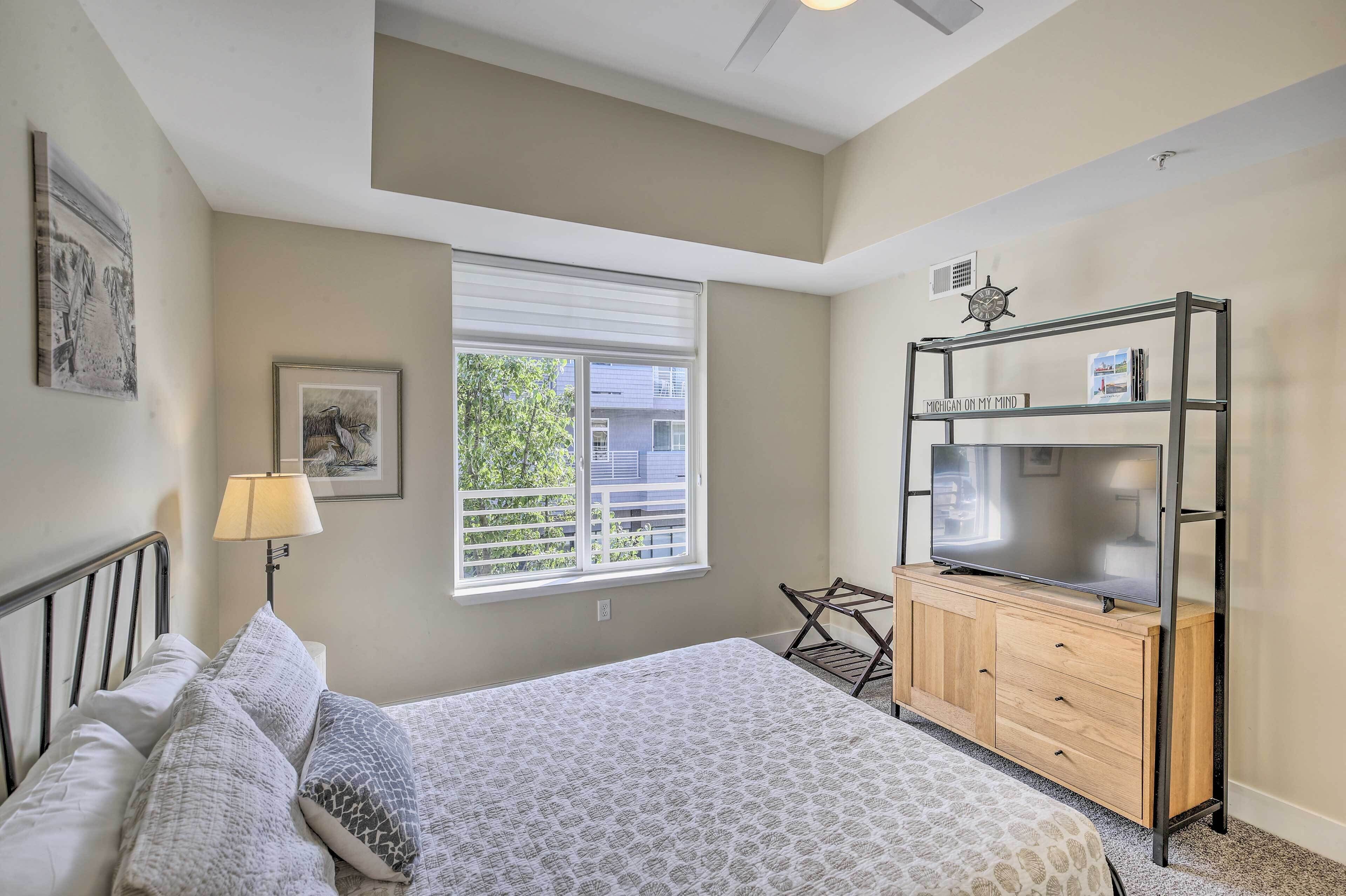A bedroom with a bed, a nightstand, a lamp, a window, a TV on a wooden cabinet, and a wall clock.