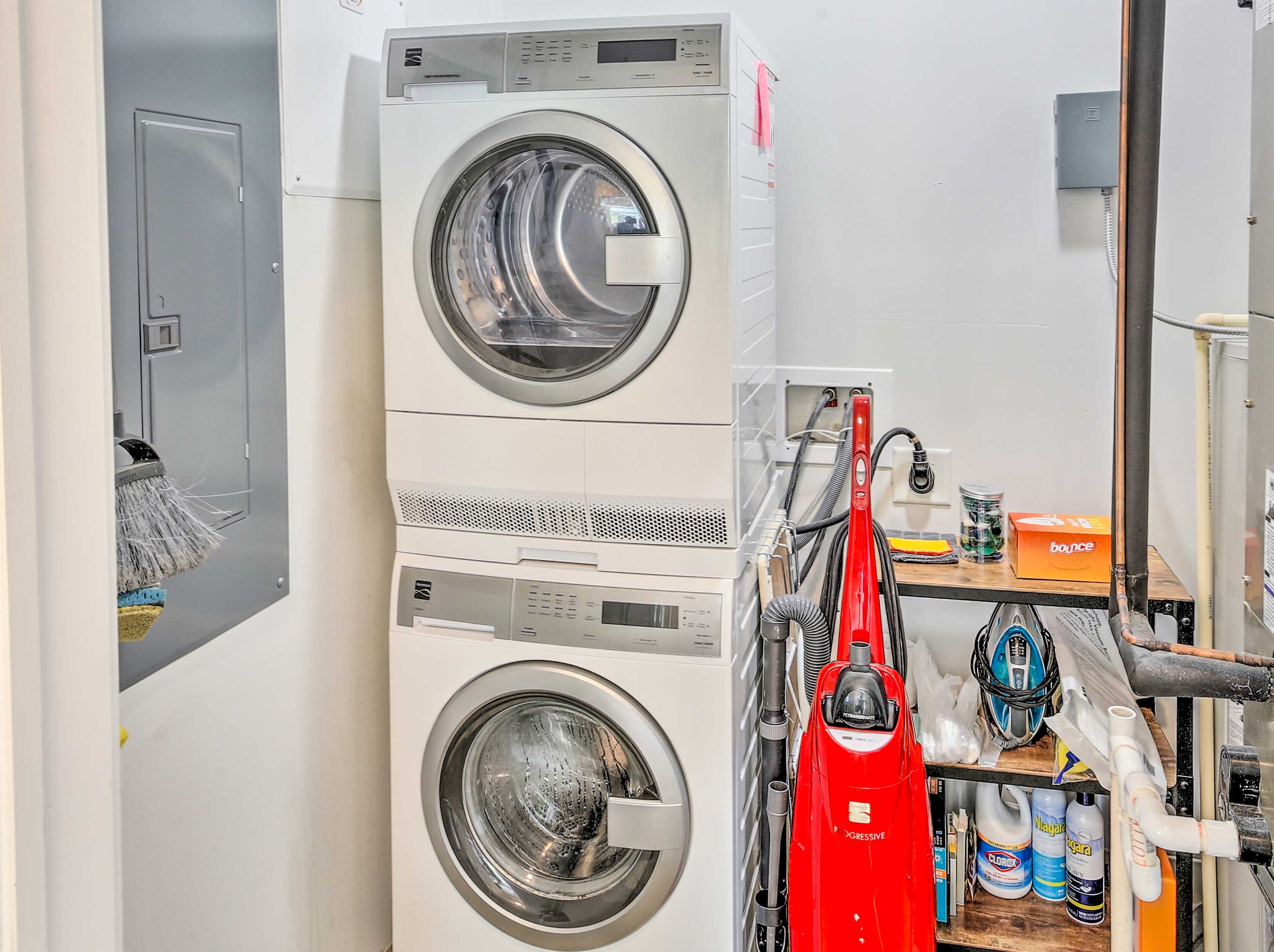 A laundry area with two stacked washing machines, a red vacuum cleaner, and various cleaning supplies on a shelf.