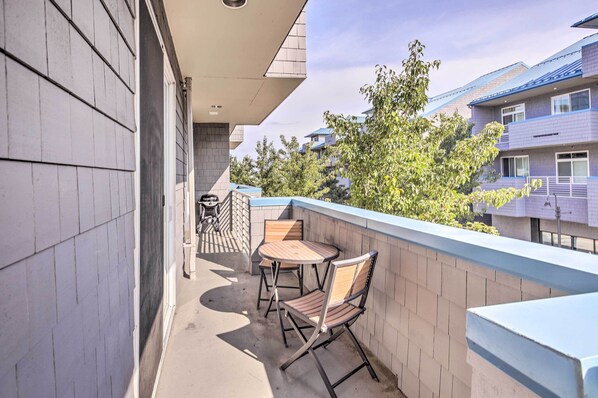 A balcony with a table and two chairs, a brick wall, and a view of neighboring buildings and trees.