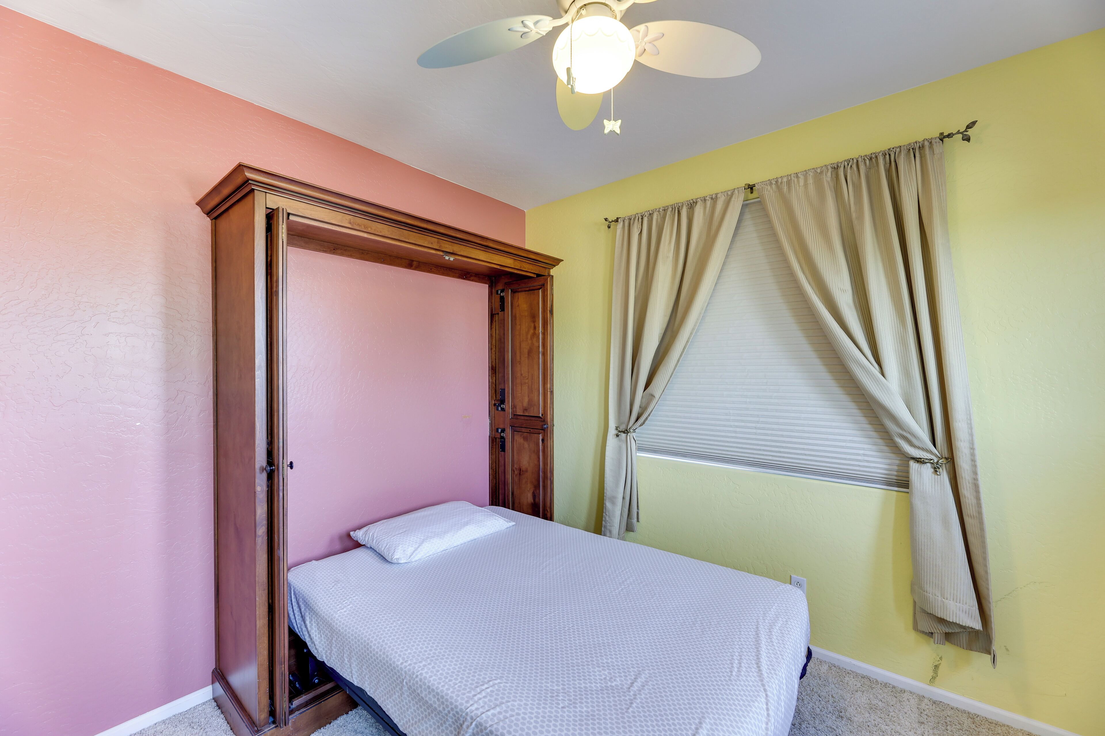 Um quarto com cama, guarda-roupa de madeira, ventilador de teto e janela com cortinas.