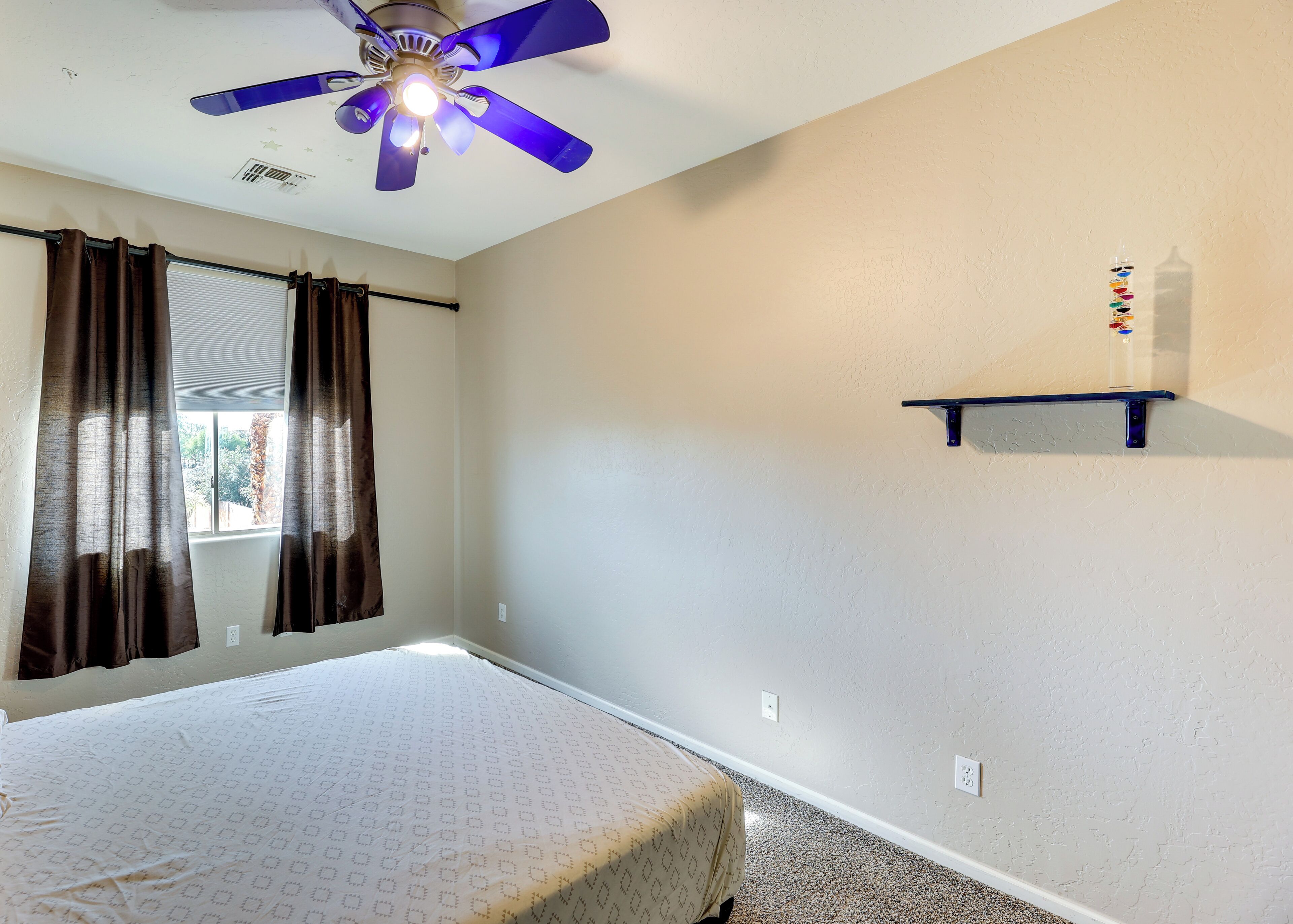 Ein Schlafzimmer mit einem Bett, einem Deckenventilator, einem Fenster mit Vorhängen und einem an der Wand befestigten Regal.