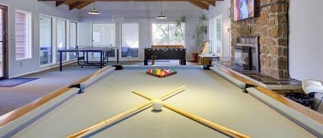 A pool table with a foosball table and a fireplace in a room with wooden beams and large windows.