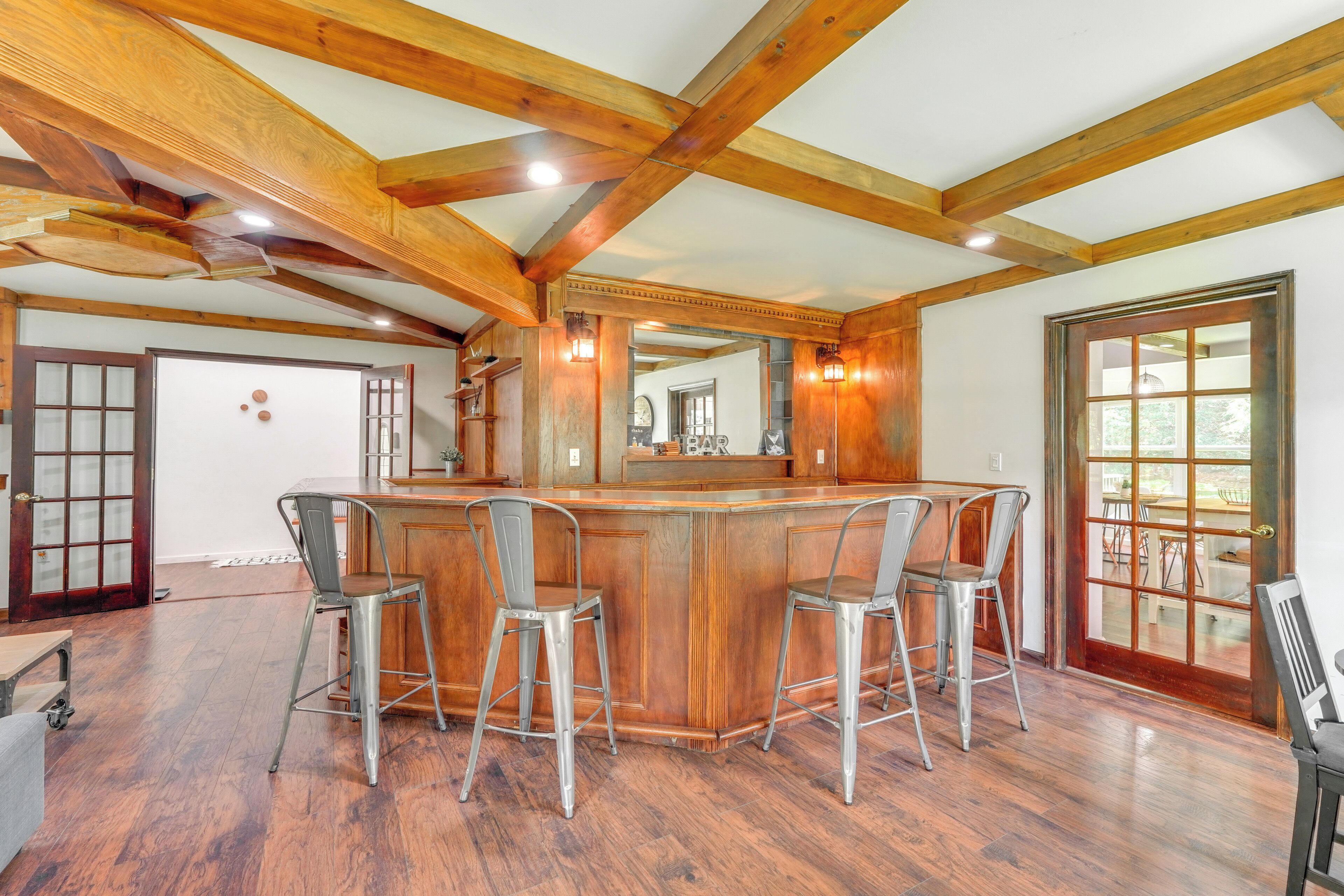 A spacious room with a wooden bar and four bar stools. The room has wooden beams on the ceiling and a large window with a view of the outdoors.