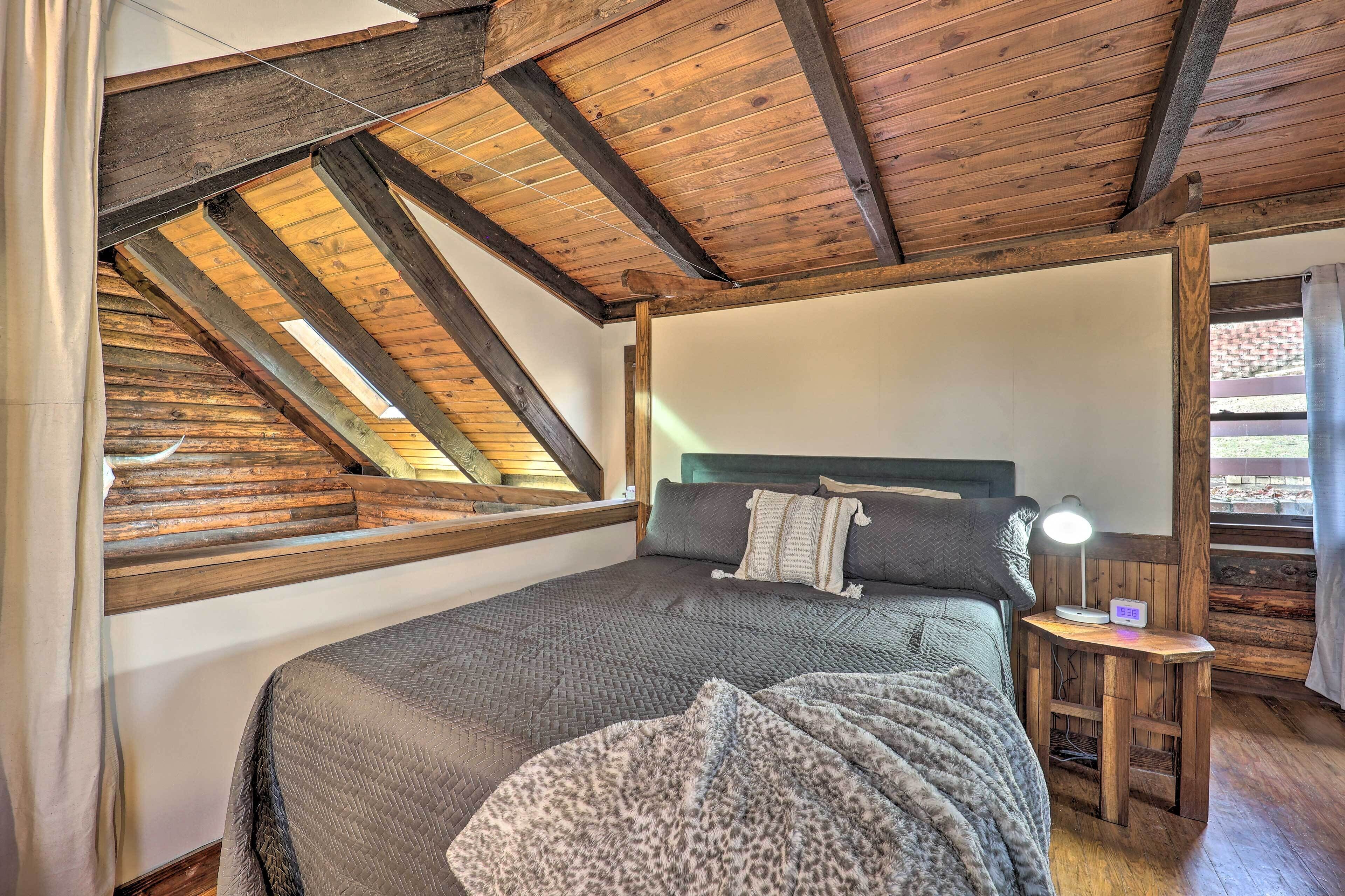 A bedroom with a sloped ceiling, a bed with a headboard, a bedside table with a lamp, and a window with a view of the outside.