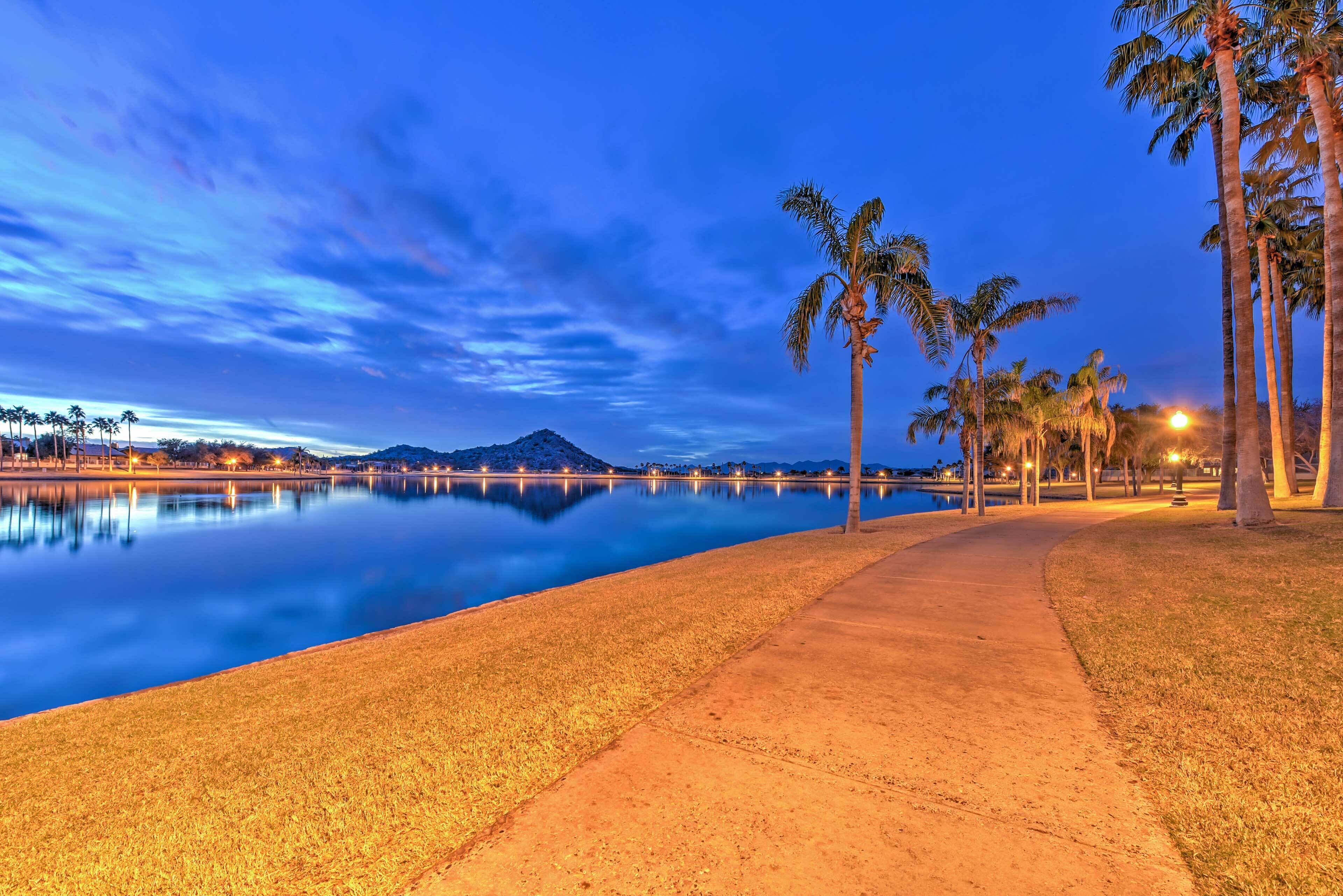 A tranquil lakeside walkway with palm trees and a mountain in the background.