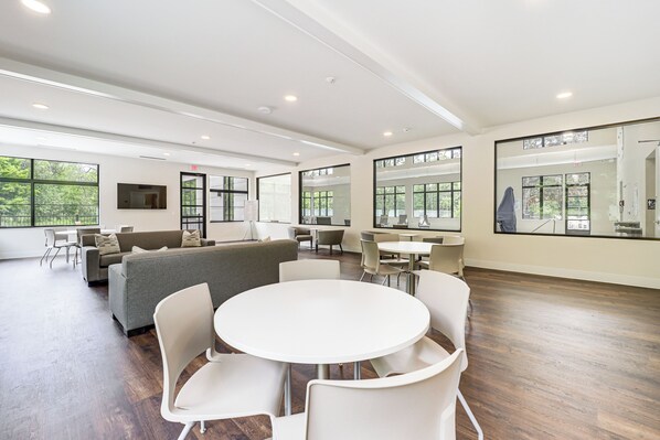 A spacious, modern room with a large round table and white chairs, a grey sofa, and a TV mounted on the wall.