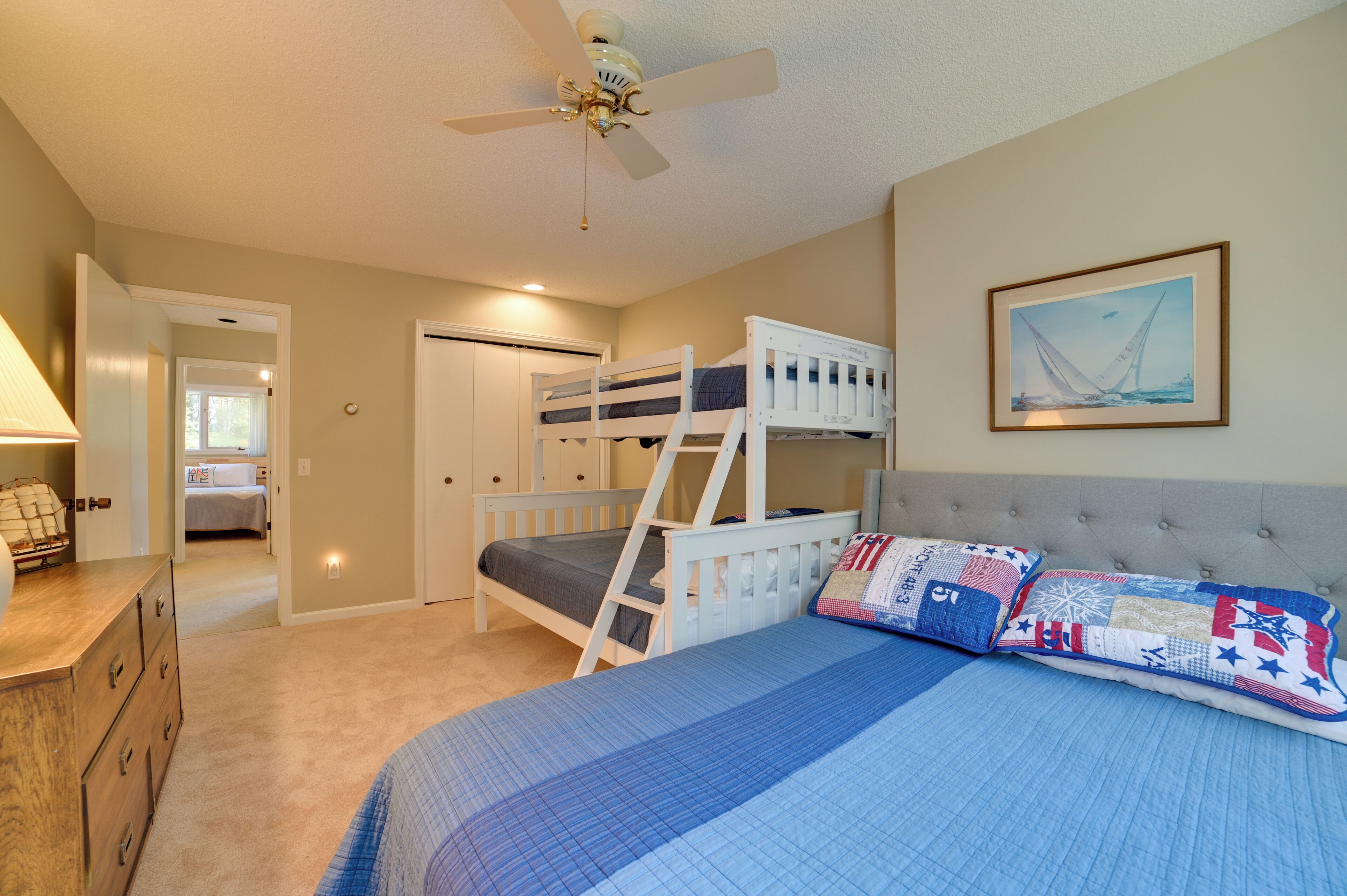 A bedroom with bunk beds, a ceiling fan, a bed with a blue quilt, a dresser, and a framed picture on the wall.