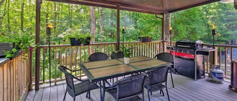 A covered outdoor deck with a table and chairs, surrounded by potted plants and a barbecue grill.